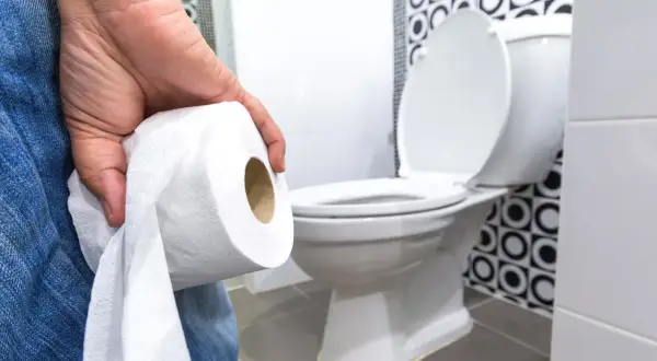 Dłoń trzymająca rolkę papieru toaletowego, w tle biała muszla klozetowa. Ktoś zmaga się z problemami z wypróżnianiem.