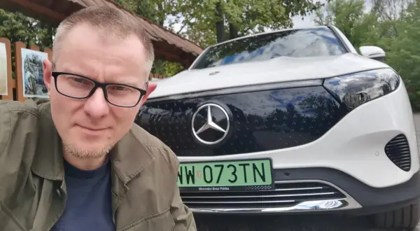 Mężczyzna w okularach pozuje do selfie na tle białego Mercedesa EQB.