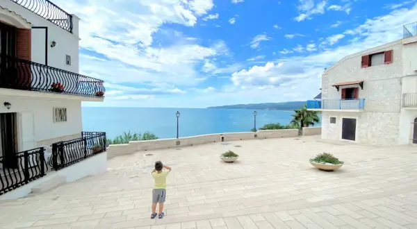 Bambino ammira il mare azzurro dalla piazzetta Petrone a Vieste, con case bianche e cielo nuvoloso.