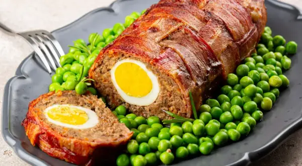 Pyszny klops z jajkiem w środku, owinięty boczkiem, podany z zielonym groszkiem.