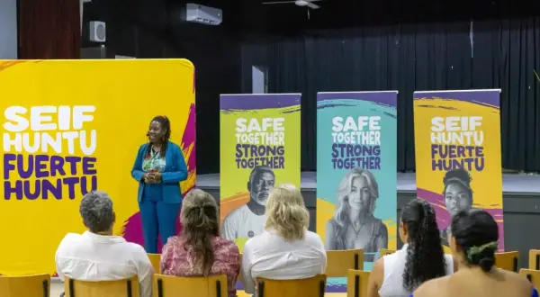 Een spreker staat voor een gele banner met "SEIF HUNTU FUERTE HUNTU". Achter haar staan banners met "SAFE TOGETHER STRONG TOGETHER". Dit is een evenement voor hss sw jeugd.