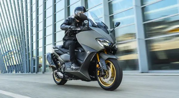 Hombre en moto clásica moderna, con chaqueta de cuero, circulando frente a un edificio de cristal.