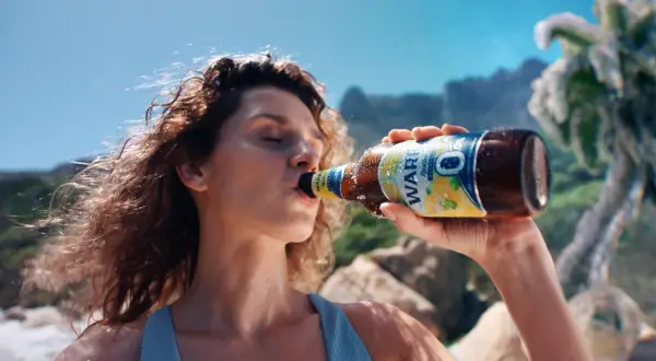 Kobieta pije piwo Żywiec 0%. Na plaży, w tle góry i palmy.