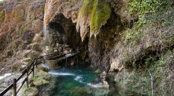 Cascata che scende tra rocce muschiose, formando una pozza d'acqua cristallina. Un sentiero con staccionata in legno conduce alle grotte di Labante.