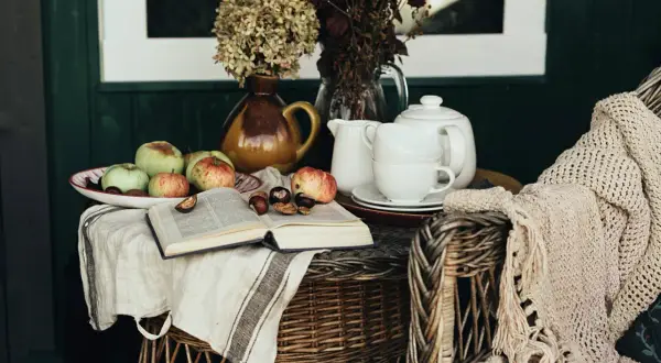 A cozy scene with apples, chestnuts, and an open book on a linen cloth, embodying the cottagecore aesthetic. A teapot and cups sit nearby, with a knitted blanket draped over a wicker chair.