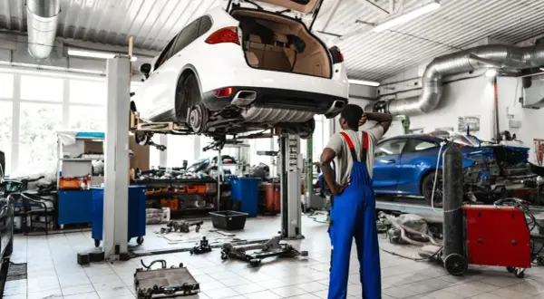 Mechanik zastanawia się, ile kosztuje założenie warsztatu samochodowego, patrząc na podniesione auto i części na podłodze.
