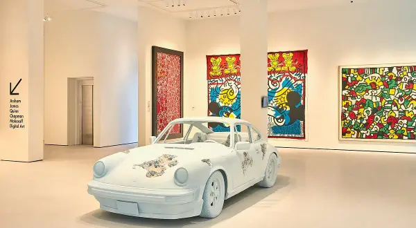 A white Porsche, artfully distressed, sits in a gallery. Vibrant Keith Haring-esque artworks adorn the walls, making for excellent Moco Museum reviews.