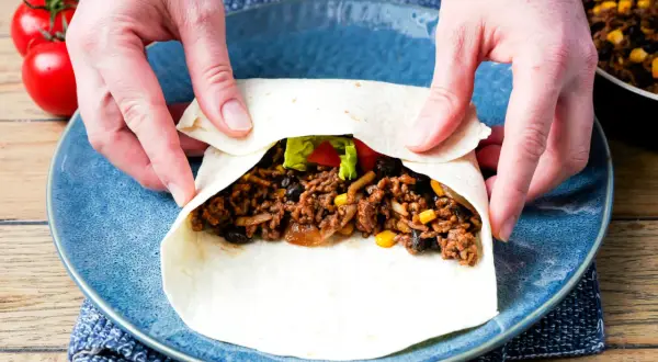 Hände falten einen Tortilla für ein leckeres burrito rezept mit Hackfleisch, Bohnen und Mais.