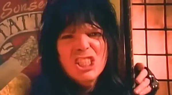 Rockman z lat 80. z burzą czarnych włosów i wykrzywioną twarzą, jakby śpiewał głośno utwory lat 80.