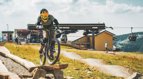 Rowerzysta pokonuje przeszkodę na drewnianej kładce w bike park Bielsko Biała. W tle kolejka linowa i budynek stacji.