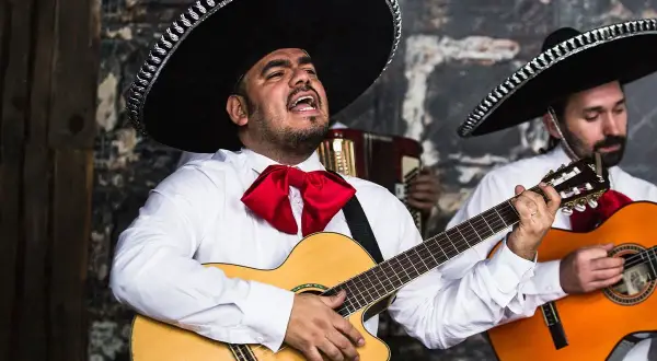 Zespół mariachi w tradycyjnych strojach, z wielkimi kapeluszami i czerwonymi muszkami, gra na gitarach i akordeonie.