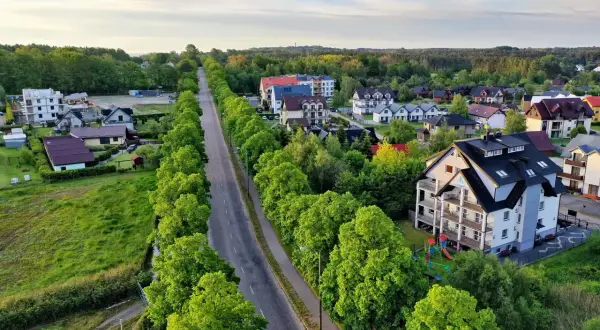 Droga w otoczeniu zieleni prowadzi do centrum Jastrzębiej Góry, gdzie widać domy i apartamentowce.