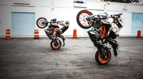 Dwóch motocyklistów wykonuje wheelie na swoich pomarańczowych motocyklach.