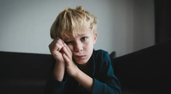 Chłopiec z blond włosami, zamyślony, opiera głowę na dłoniach. Jego spojrzenie sugeruje trudności, być może związane z zespołem Aspergera.