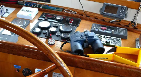 Un binocolo da ingrandimento, pronto per scrutare l'orizzonte, riposa sul cruscotto di una barca, accanto a strumenti di navigazione e a un timone in legno lucido.