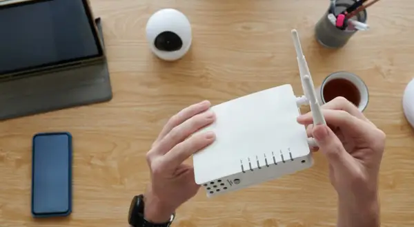 Ręce trzymają biały router, obok tablet, telefon i kubek herbaty. Czas na konfigurację sieci i hasło do sieci wifi.