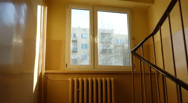 Słoneczne promienie padają na błyszczącą ścianę w klatce schodowej. Widać okno z widokiem na blok i kaloryfer.