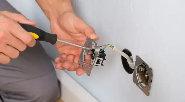 Elektryk wykonuje mostkowanie gniazdka, przykręcając mechanizm do puszki. Widać przewody elektryczne.