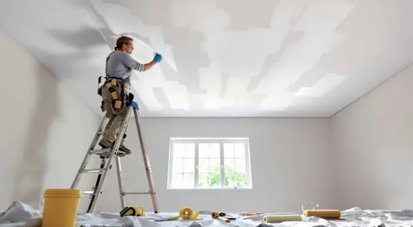 Un peintre sur une échelle applique une toile de rénovation plafond. Des rouleaux et un seau sont prêts.