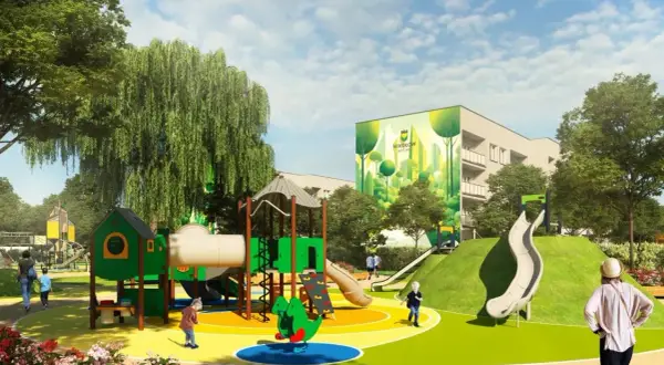 Kolorowy plac zabaw z zielonymi elementami, zjeżdżalniami i huśtawkami, na tle budynku z muralem.