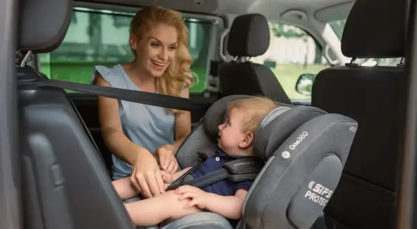 Lächelnde Mutter sichert ihr Baby im Kindersitz vorne im Auto.