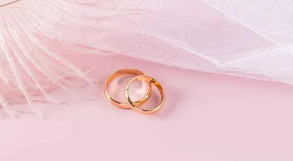 Dos anillos de oro rosa entrelazados, sobre un fondo rosa pálido con una pluma blanca y tela delicada.