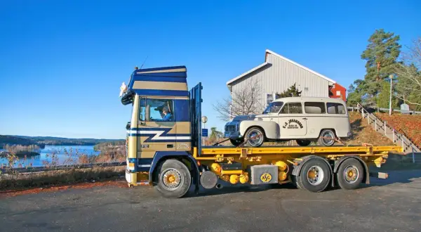 En blågul lastbil transporterar en vit Volvo Raske herrgårdsvagn längs en sjö.