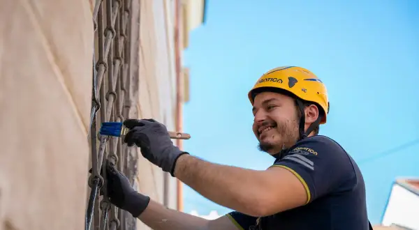 Hombre con casco amarillo sonríe mientras realiza el revoco de paredes con una brocha azul.