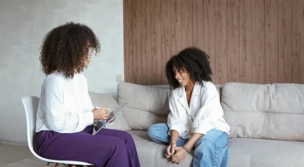Due donne parlano in un ambiente accogliente. Una, seduta su una sedia, usa un tablet, mentre l'altra è rilassata sul divano. Potrebbe essere una sessione di psicoterapia breve strategica cos'è.
