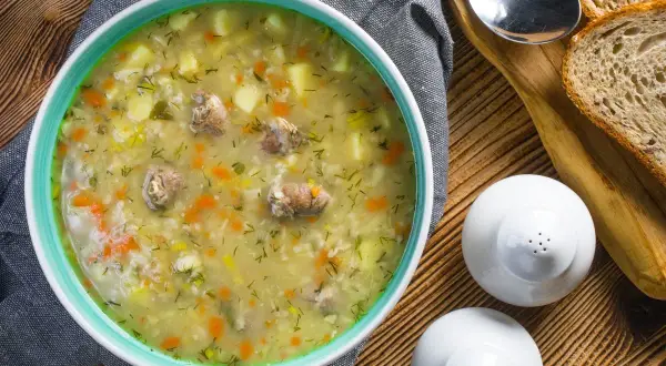 Aromaticzny krupnik, przepis babci, z kawałkami mięsa, ziemniaków i marchewki, posypany koperkiem. Obok kromki chleba i solniczki.