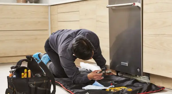 A technician kneels to replace a dishwasher, surrounded by tools and a protective mat.
