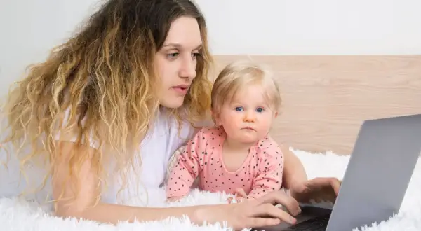 Kobieta z dzieckiem na rękach pracuje przy laptopie, zastanawiając się, czy na urlopie wychowawczym można pracować.