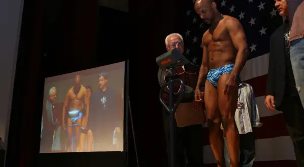 A muscular man in blue patterned shorts stands on a scale, preparing for his junior bantamweight weight check. An official looks on.