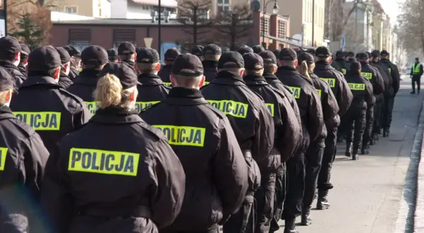 Policjanci w czarnych kurtkach z napisem "POLICJA" stoją w szeregu, gotowi do akcji. Widać, jak pracuje policja zmiany, przygotowując się do służby.