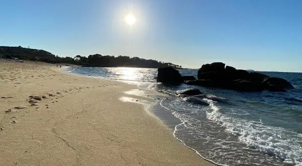 La **playa de Tulla** bajo un sol radiante. Huellas en la arena dorada y el mar azul acarician las rocas oscuras.