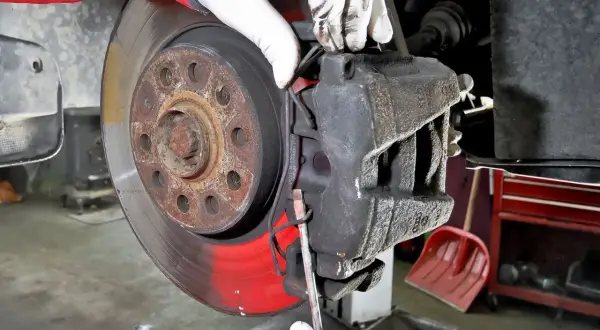 Mechanik w białych rękawiczkach dokonuje wymiany klocków hamulcowych, używając śrubokręta do podważenia starego klocka.