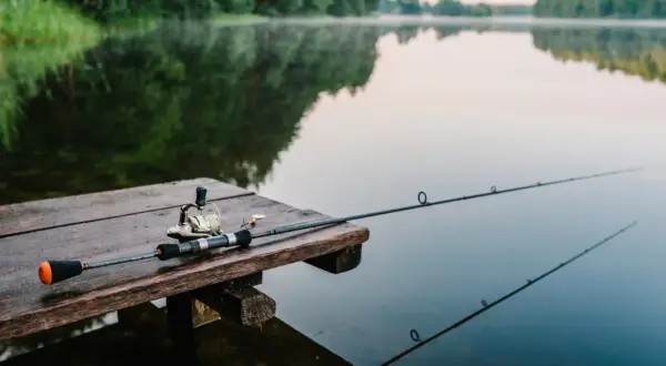 Wędka i kołowrotek leżą na drewnianym pomoście, gotowe do wędkowania na Mazurach. Spokojna tafla jeziora odbija zalesione brzegi.