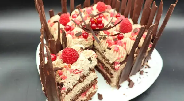 Una golosa torta cioccolato e lamponi, decorata con panna, frutti rossi e scaglie di cioccolato. Una fetta è già stata servita.
