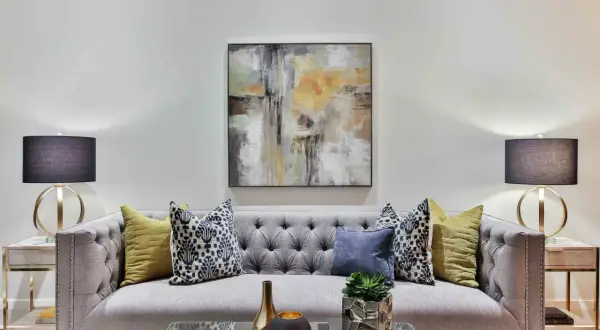 Nowoczesna szara sofa z pikowanymi guzikami, ozdobiona poduszkami i abstrakcyjnym obrazem. To idealne miejsce na relaks, tworzące przytulną atmosferę.