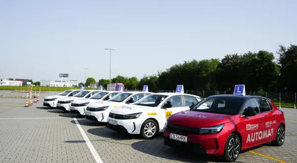 Rząd białych samochodów nauki jazdy z literą "L" na dachu i czerwony Opel z napisem "AUTOMAT" na placu manewrowym. Sprawdź, jaki jest prawo jazdy b+e koszt.