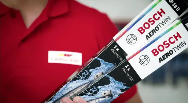 Person i röd pikétröja med Carglass-logga håller upp två paket med Bosch Aerotwin torkarblad.