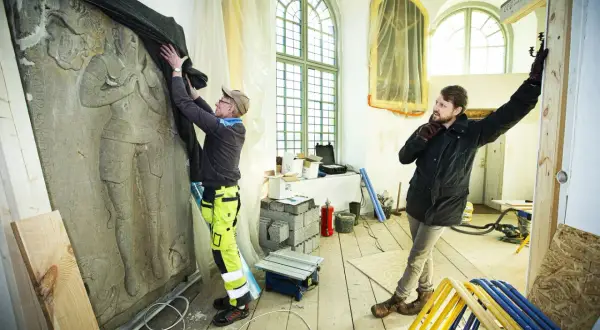 Två män arbetar med en stor stenrelief i en halvrund del av kyrka under renovering.