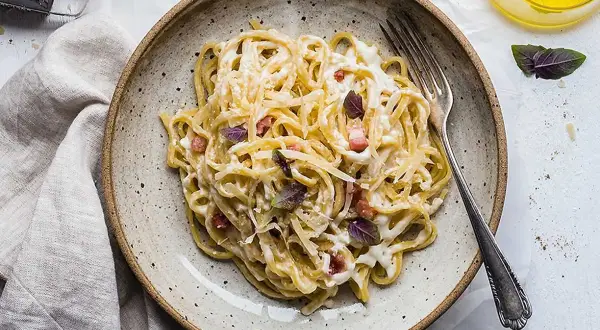 En tallrik med krämig, klassisk carbonara, toppad med basilika och parmesan. En gaffel ligger redo att avnjutas.