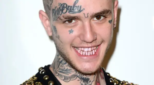 Uśmiechnięty Lil Peep z tatuażami na twarzy i szyi, błyszczącymi różowymi zębami. Pytanie czy lil peep żyje, pozostaje w sercach fanów.