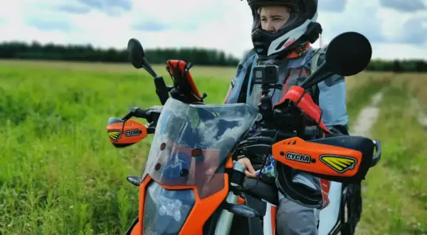 Motocyklista w kasku i kurtce przygotowuje się do podróży motocyklem po Europie. W tle zielone pole i niebo.