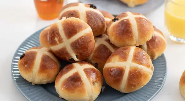 En hög med gyllene, nybakade **hot cross buns** på en blå tallrik, redo att avnjutas med honung och apelsinjuice.