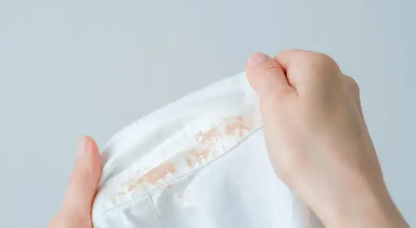 Dłonie trzymają białą koszulę z widocznymi plamami. Dowiedz się, jak usunąć plamy z podkładu, aby przywrócić ubraniu nieskazitelny wygląd.