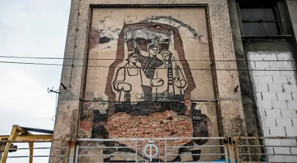 Zniszczony mural w stylu socrealizmu przedstawiający trzech robotników z karabinami.