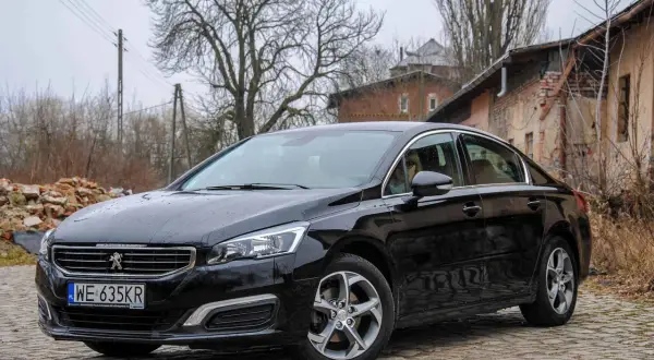 Czarny Peugeot 508 na brukowanej drodze. W tle zrujnowane budynki i drzewa. Wiele osób szuka peugeot 508 opinie przed zakupem.