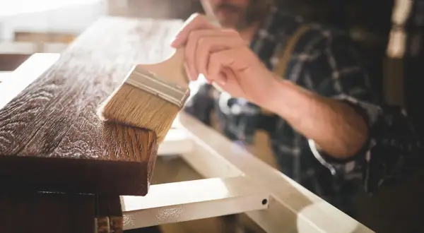 Un falegname applica una finitura protettiva su un pezzo di legno. Dopo l'impregnante, cosa si mette? Una vernice trasparente o una cera per esaltare la venatura.